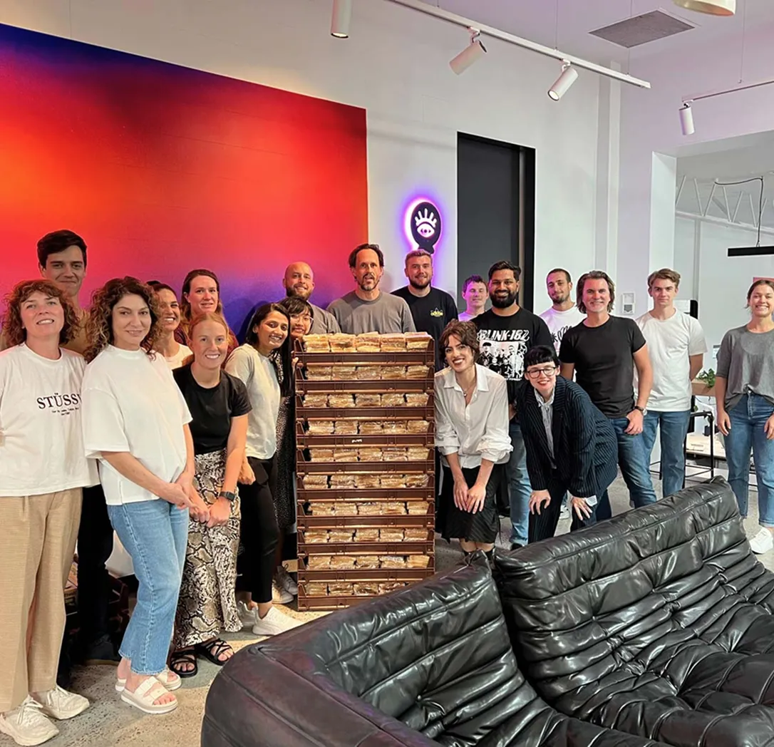 A group of about 18 people stand and smile around a tall stack of boxed pastries in a modern, colourful office space with sofas and vibrant wall art.