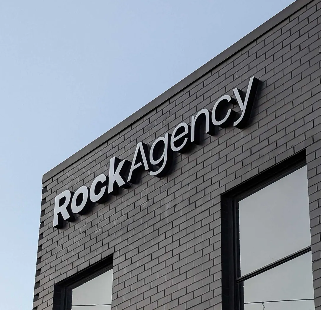 A modern building with dark grey brick exterior features a large RockAgency sign in bold, black letters against a clear blue sky. Two large windows are visible below the sign.