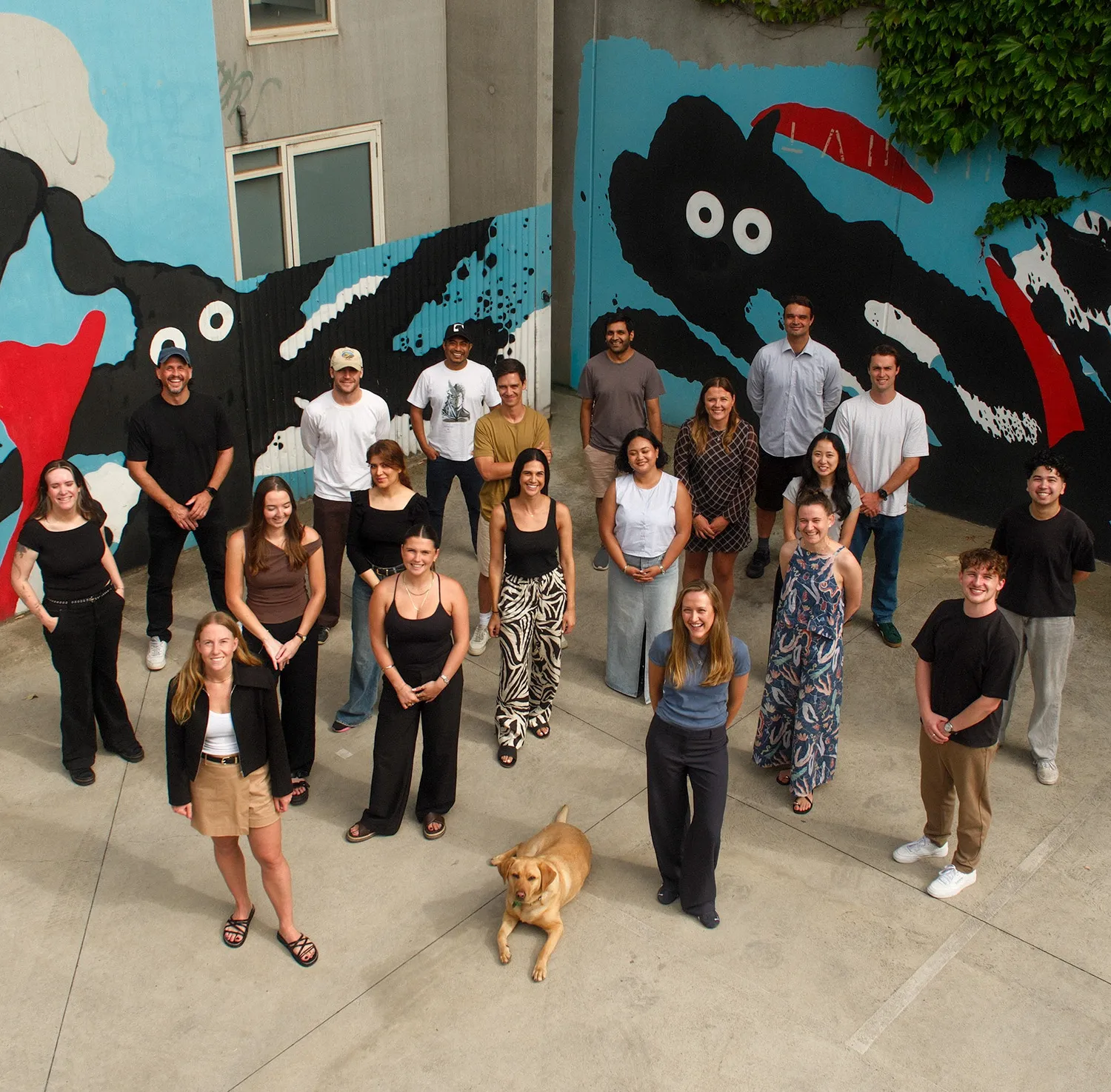 A group of people and a yellow dog stand outdoors on concrete in front of a colourful mural with abstract shapes and eyes, smiling and facing the camera.