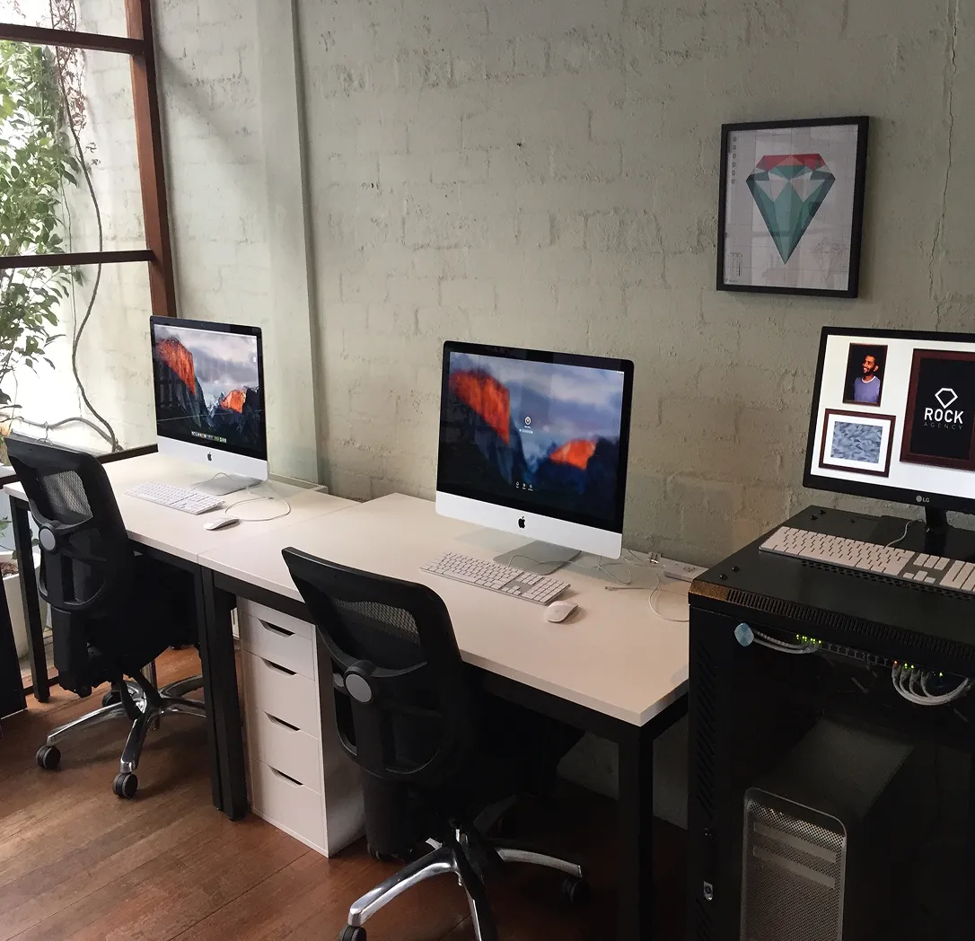 A modern office workspace with three iMac computers on white desks, black office chairs, a framed geometric art on the wall, and greenery visible through a window on the left.