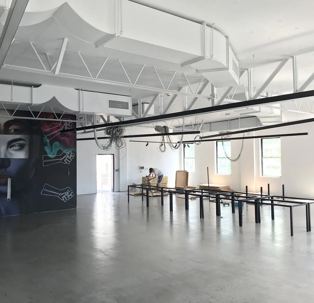 A spacious industrial-style room with exposed beams, large windows, and unfinished furniture. A person works at a table, and a colourful mural of a woman’s face decorates one wall. Natural light fills the space.